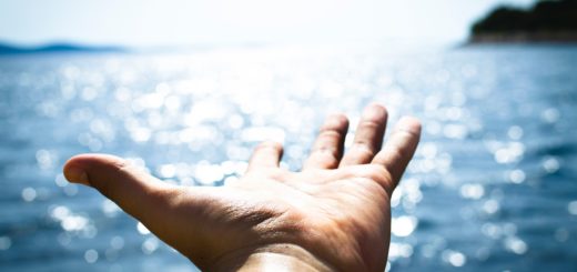 Hand reaching out over a serene body of water with sunlight reflecting on the surface.