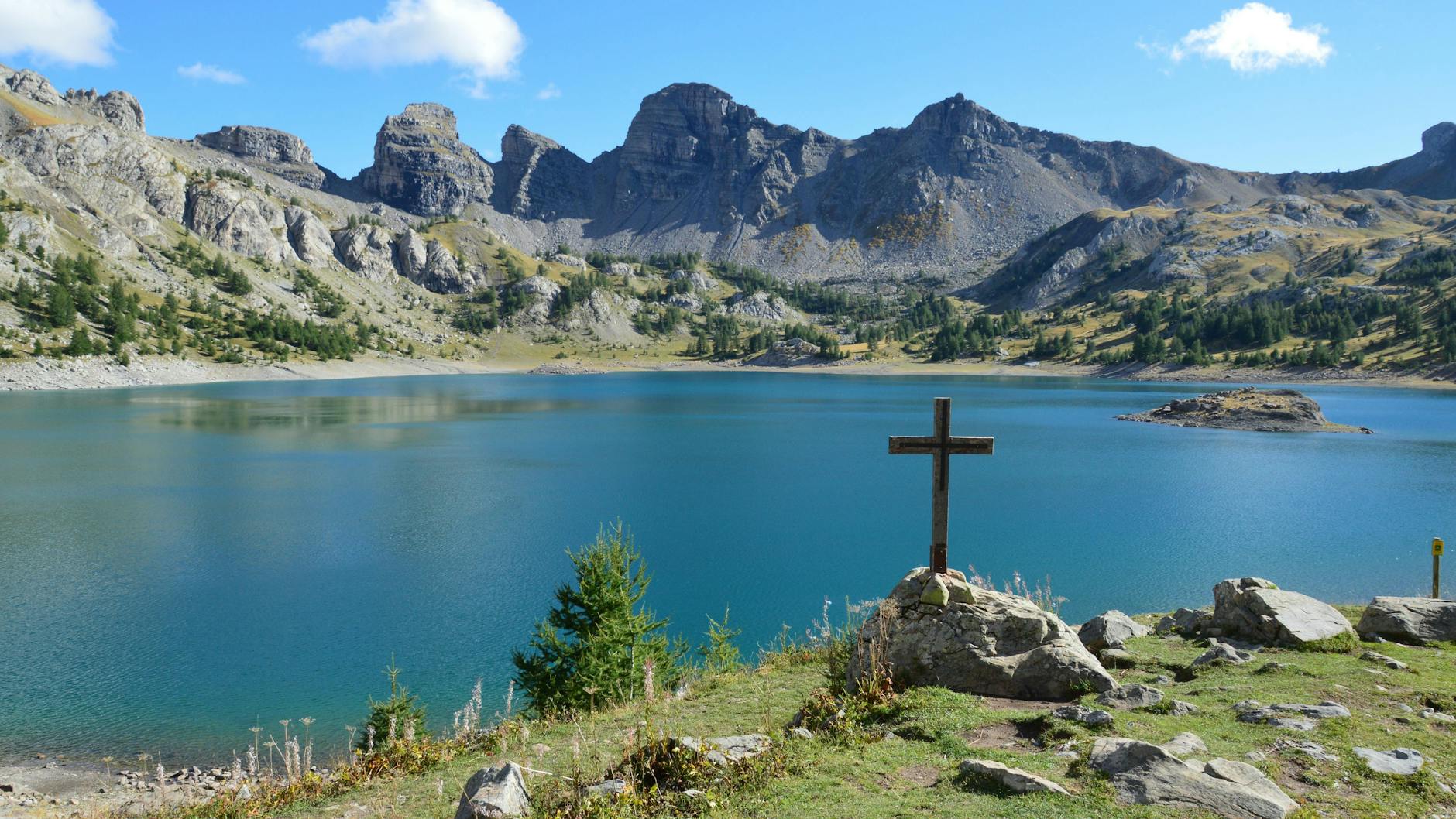 Serene mountain lake with a wooden cross on the rocky shoreline and lush greenery, symbolizing faith and spirituality.